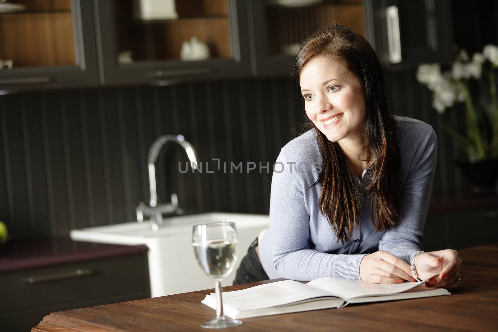 Attractive young woman reading a recipe from a cookery book in her kitchen with a glass of wine.