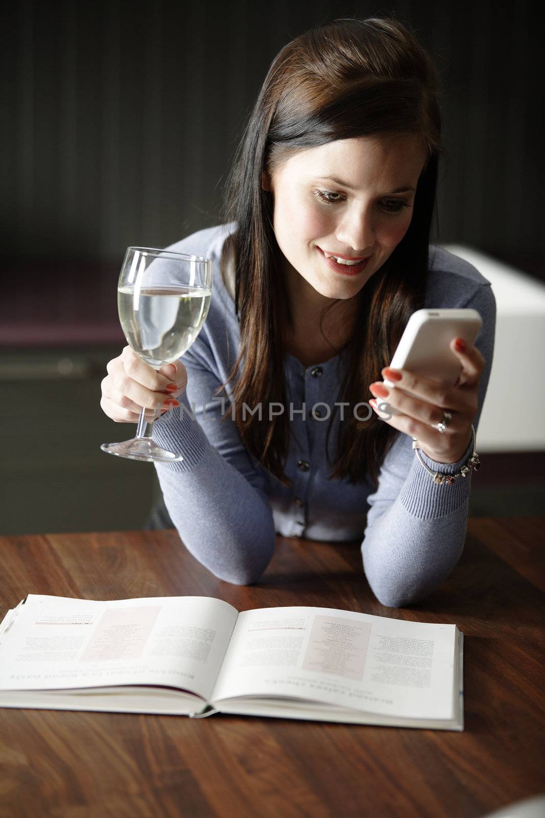 Attractive young woman enjoying a glass of wine in her kitchen while texting on the phone.