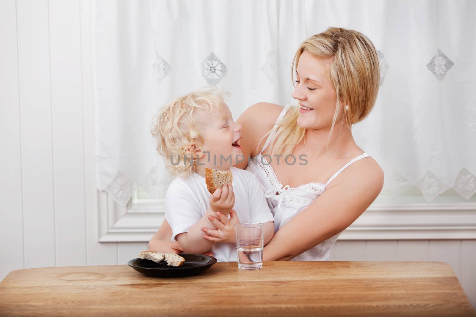 Young beautiful mother looking at cheerful baby boy eating bread at dining table