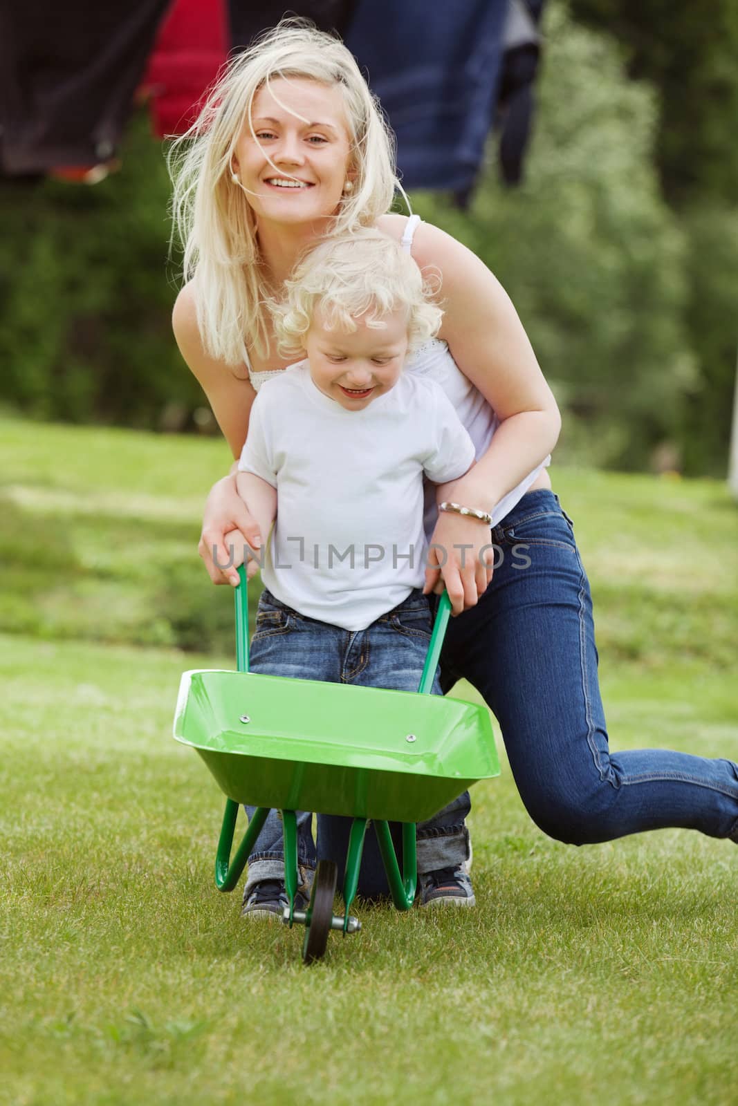 Portrait of young woman helping her toddler boy push a wheelbarrow in garden