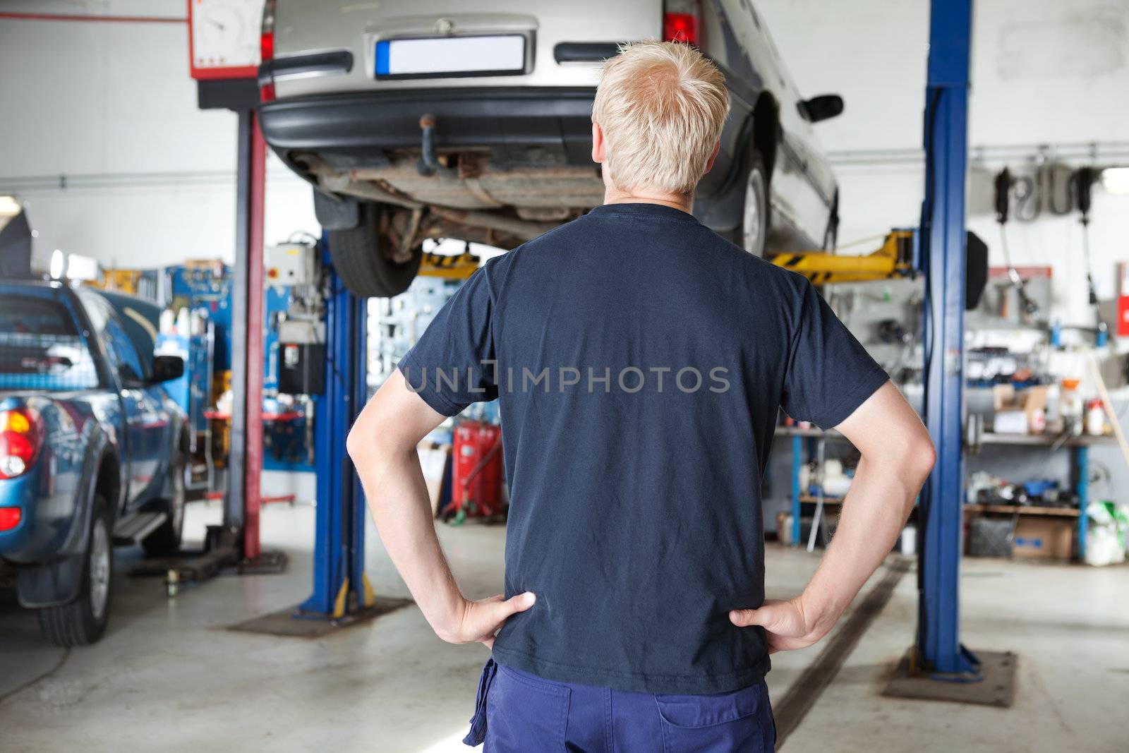 Rear view of car repairer looking at car in auto repair shop with hands on waist