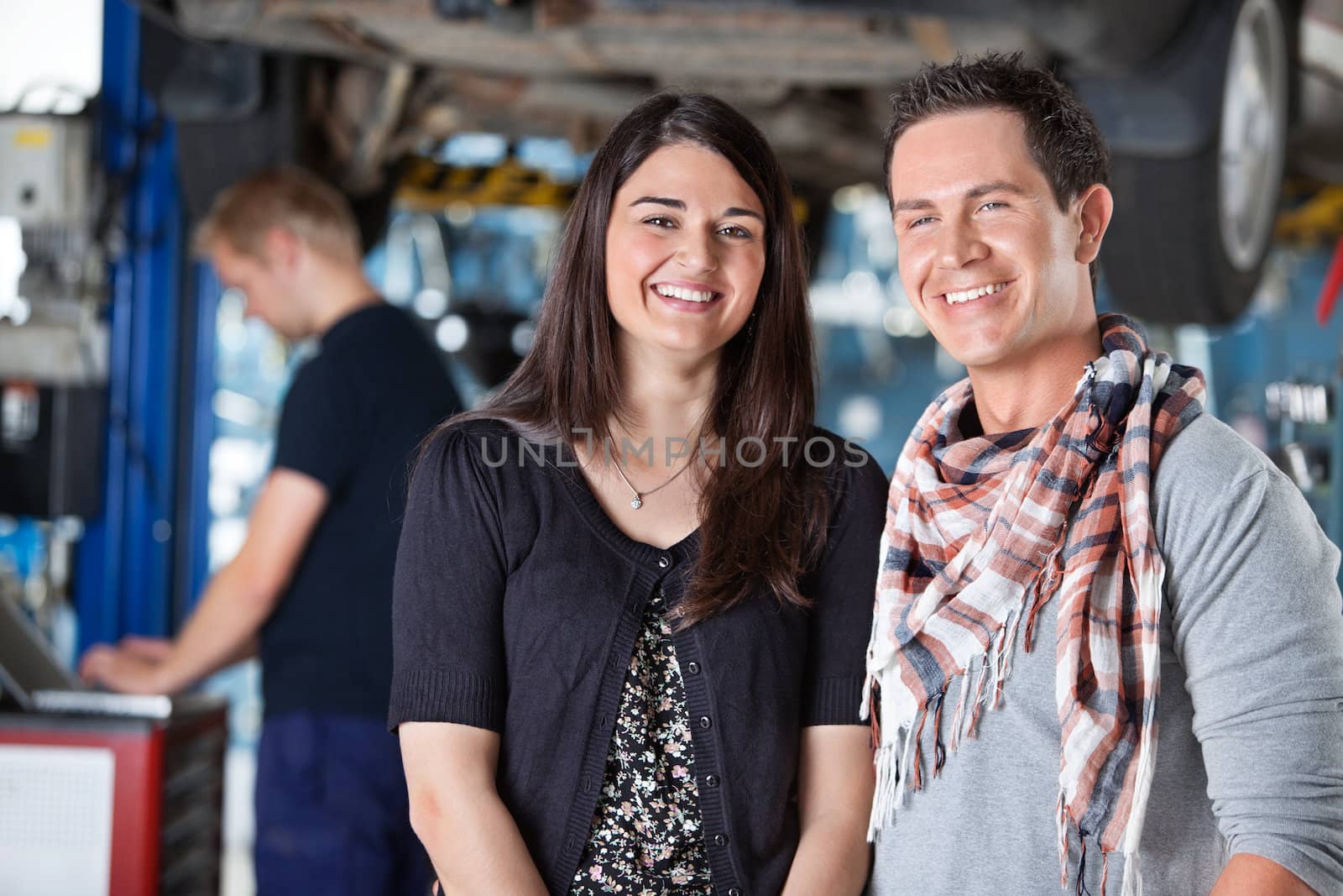 Portrait of smiling young couple in auto repair shop with mechanic in the background