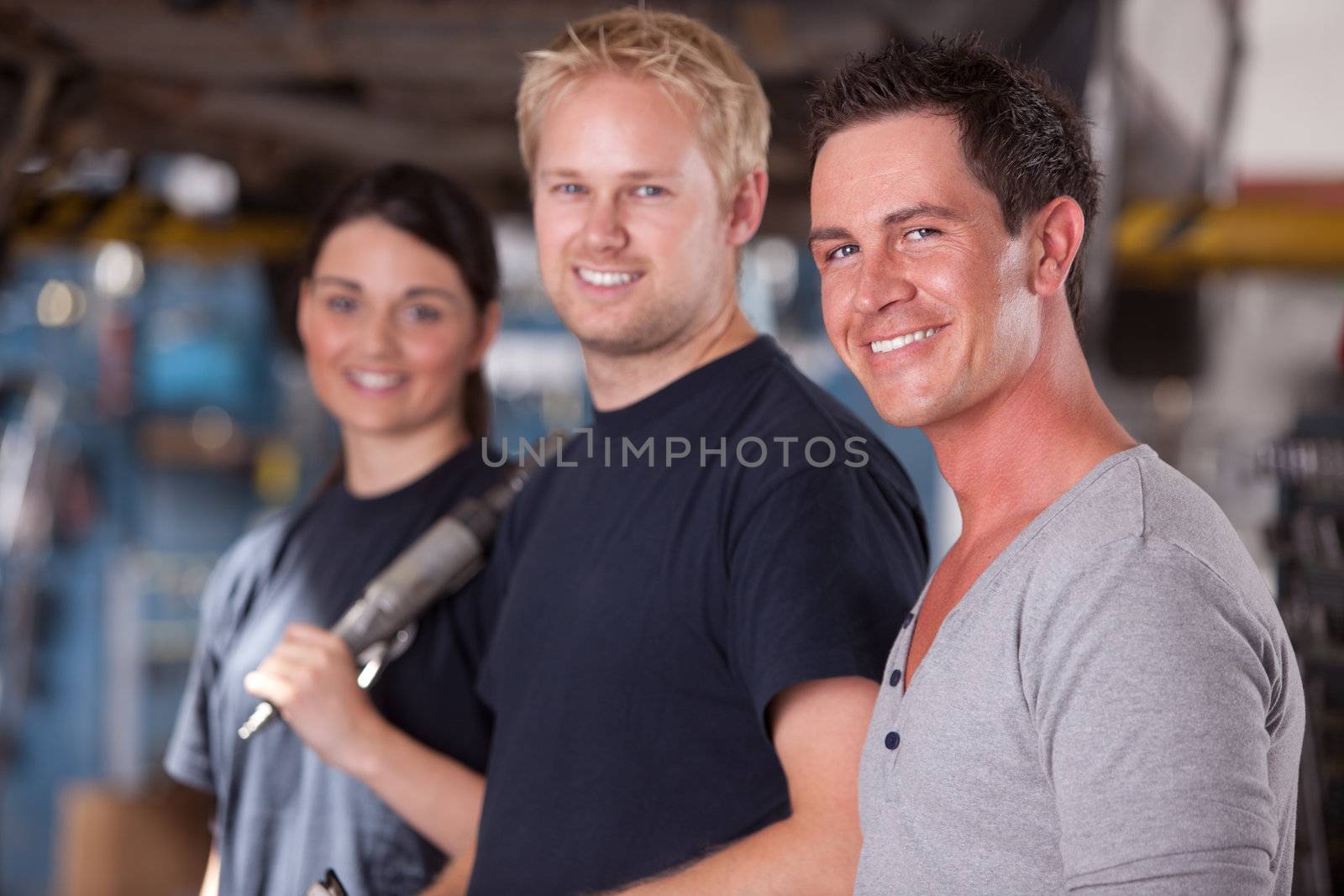 A group of three mechanics in a auto repair shop, looking at the camera