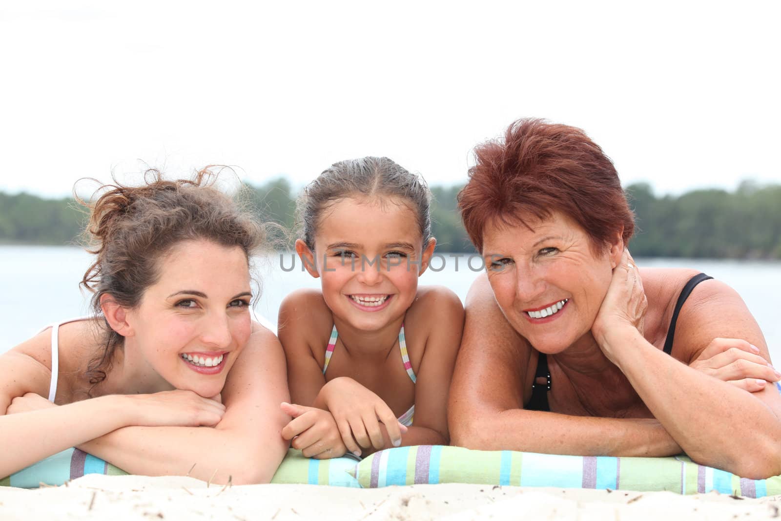 a 30 years old woman, a little girl and a 55 years old woman lying down on the beach, behind sea and forest background by phovoir