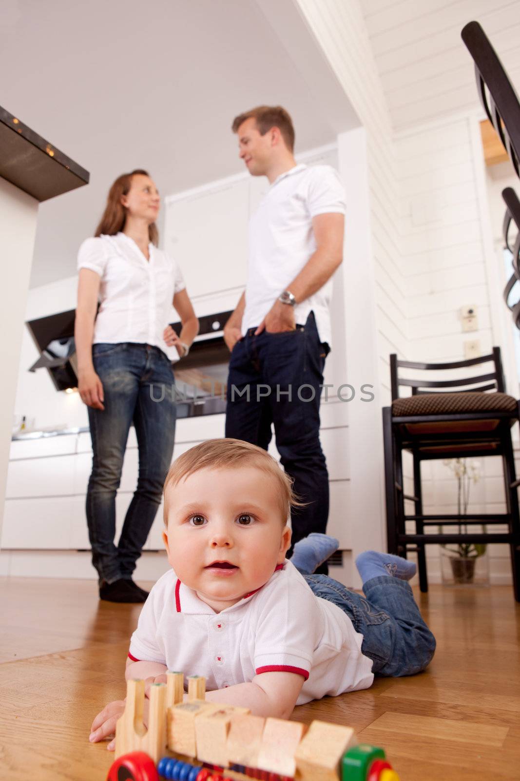 A happy child playing on kitchen floor with parents talking in the background