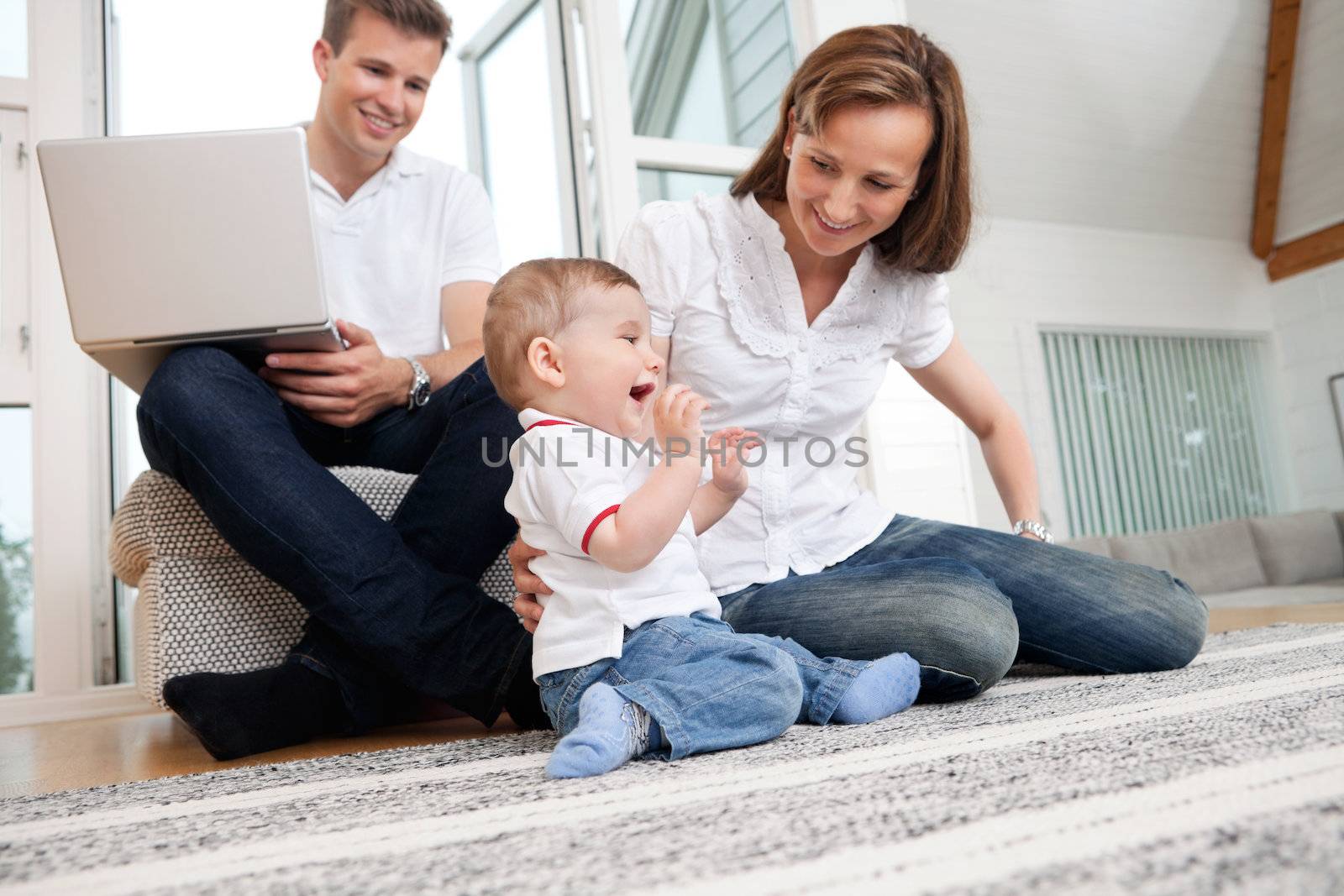 Smiling mother playing with cute child with man using laptop in background