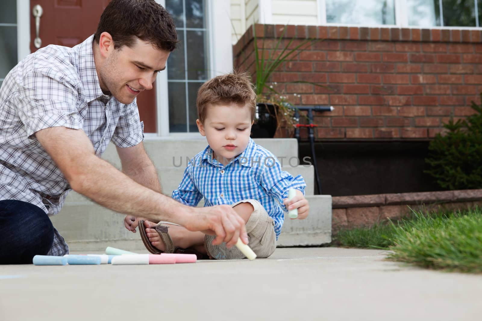 Young father drawing with chalk on the ground with son sitting beside