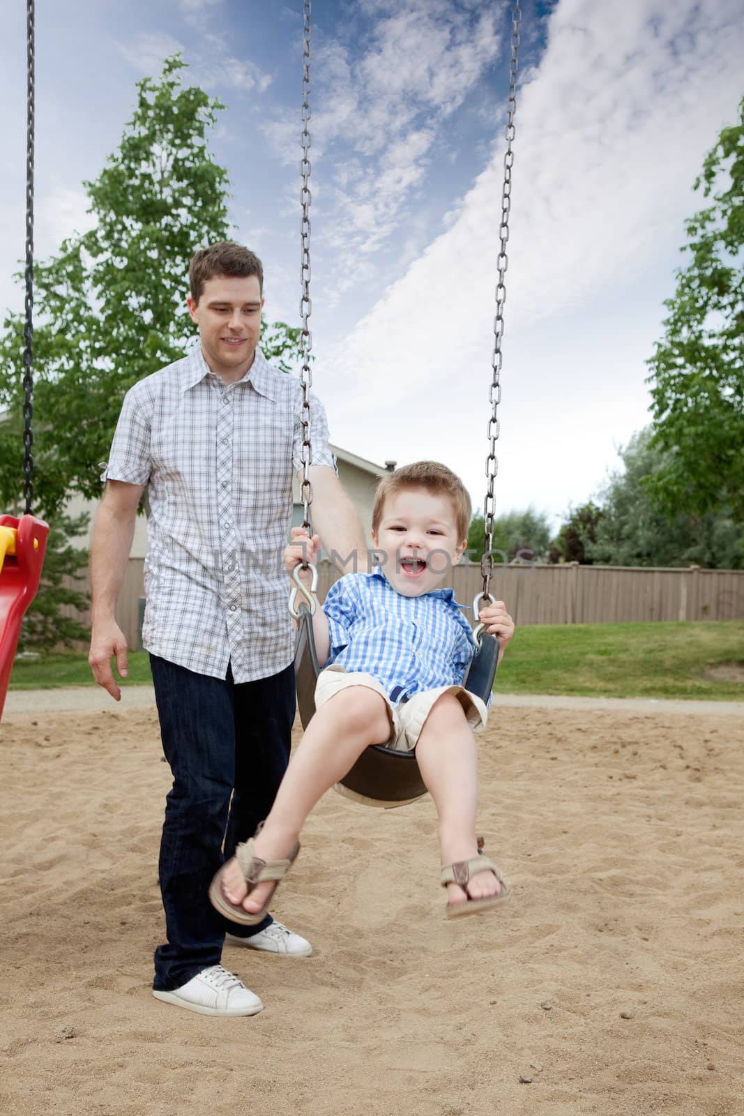 Happy Father Pushing Boy On Swing In Playground.
