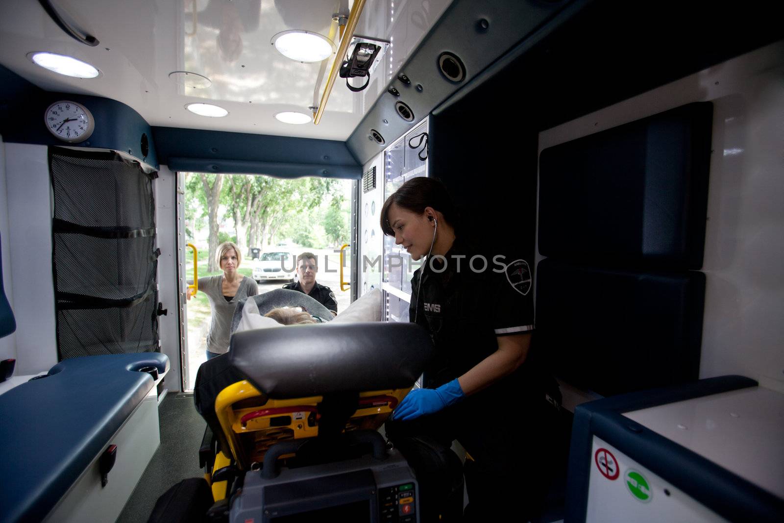 Paramedic tending to an ill patient with ambulance driver and caregiver looking in from rear doors