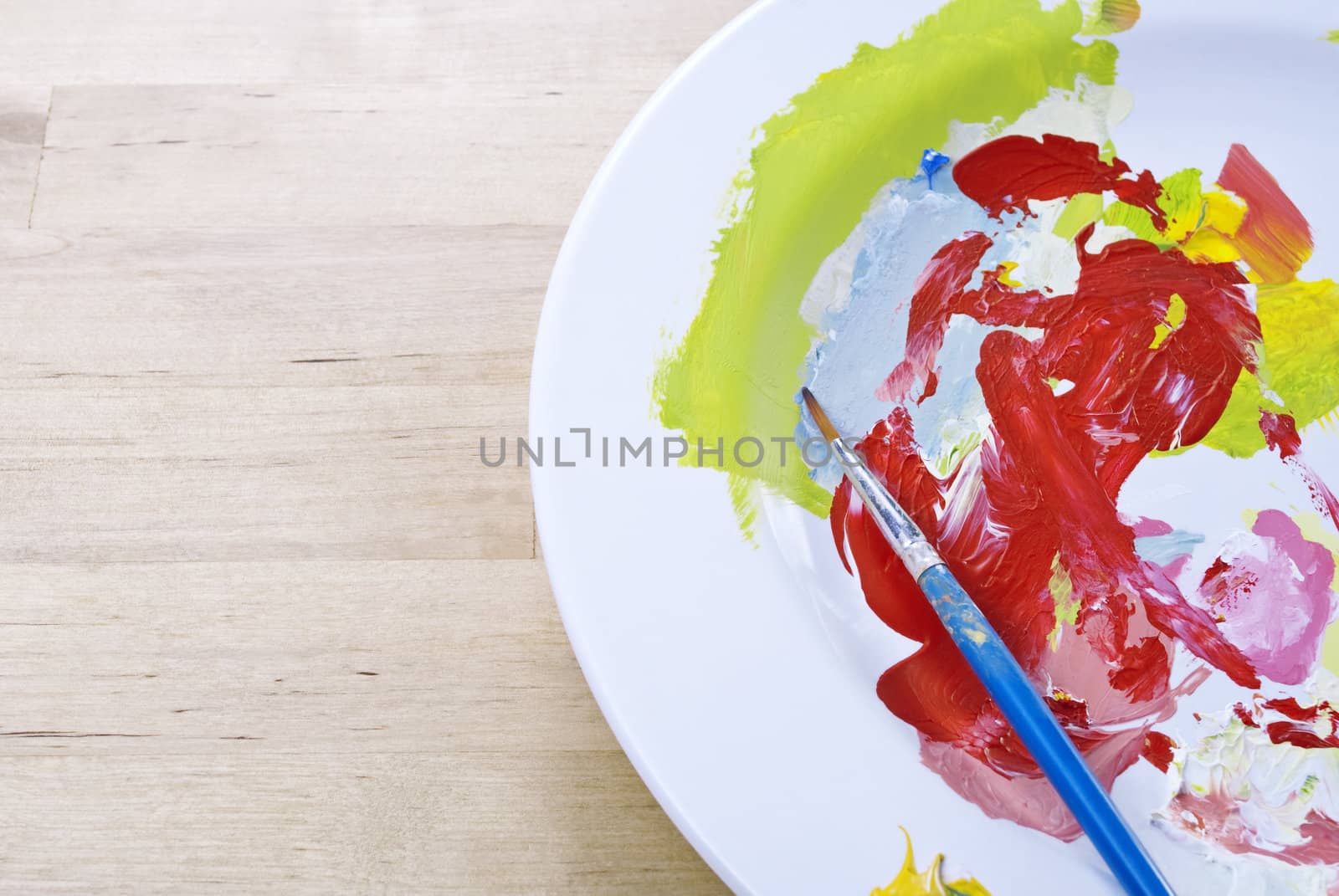 Colourful paints mixed on a china plate palette on a birch wooden table. Paintbrush resting on plate.  Copy space on left hand side.