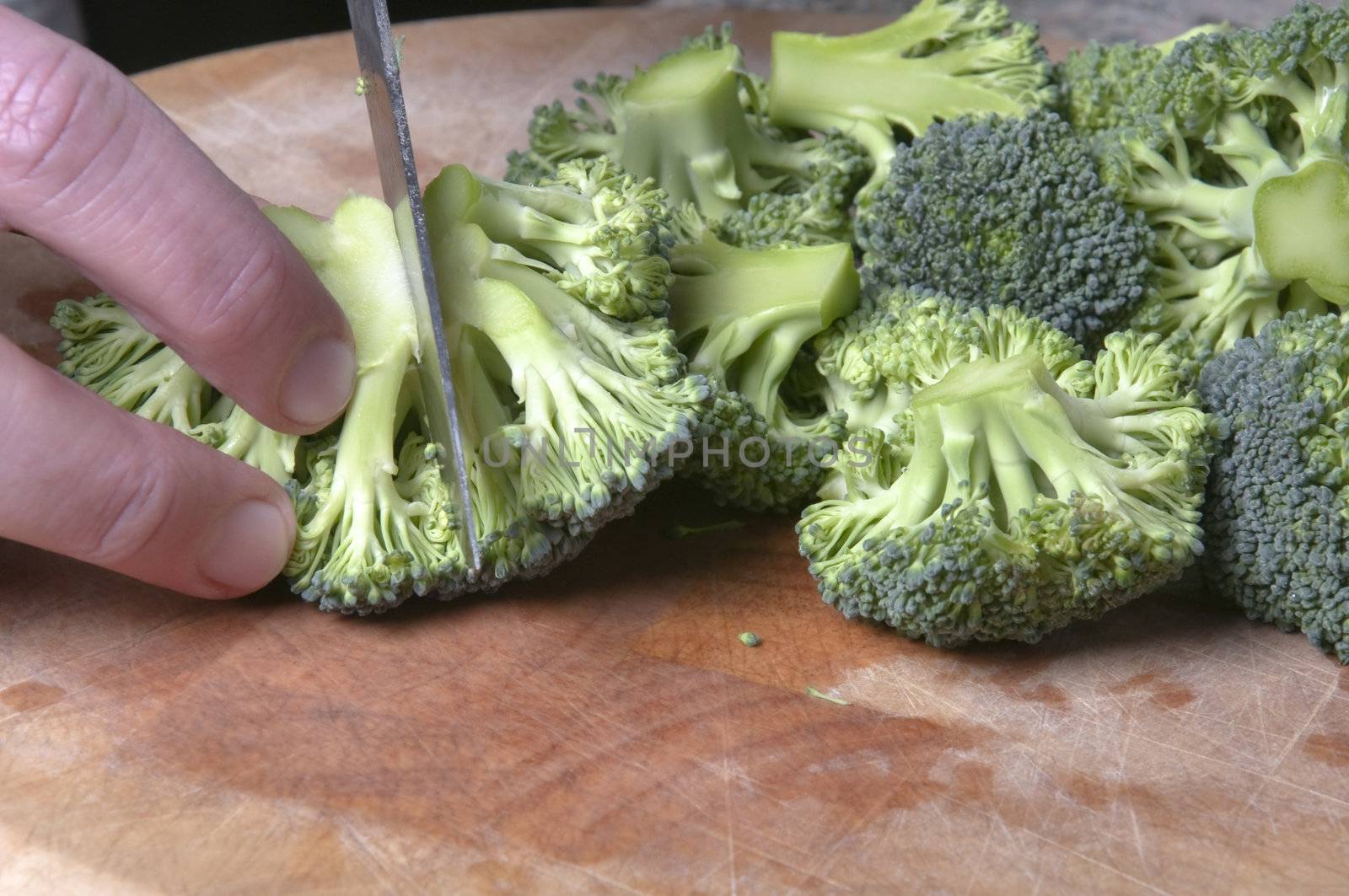 Close up of a man's hand chopping washed broccoli with a steel knife on a wooden chopping board.  Some small water droplets visible.