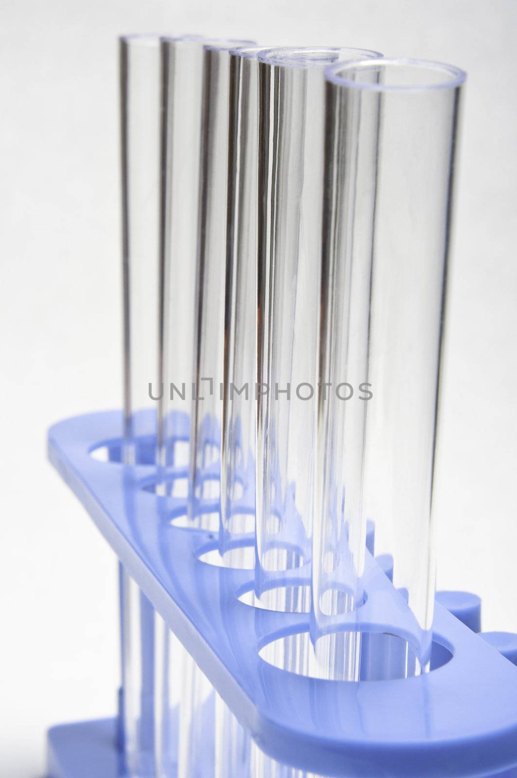 Macro shot of six test tubes, held upright in a blue stand.  Slightly angled and leading away from the viewer.  Soft white background.