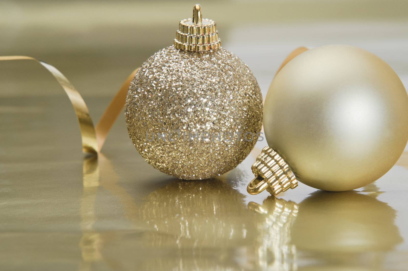 Two gold Christmas baubles to right of frame with spiralling foil ribbon in the background.  Set on a gold reflective metallic surface providing copy space below.