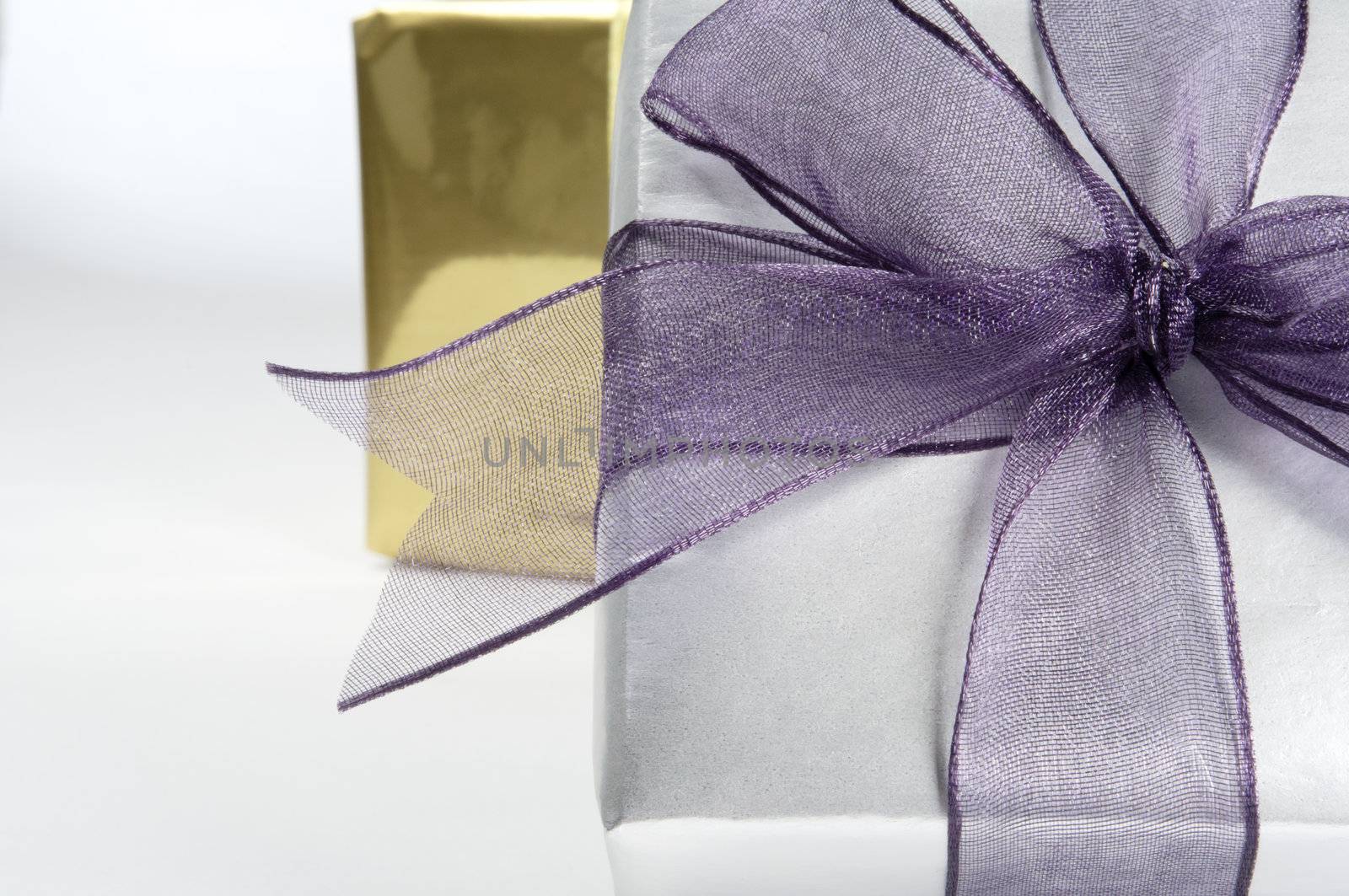 Close up (macro) of a gift box wrapped in silver paper with purple organza ribbon tied in a bow.  A further gift wrapped in gold in soft focus background.  Copy space to left.