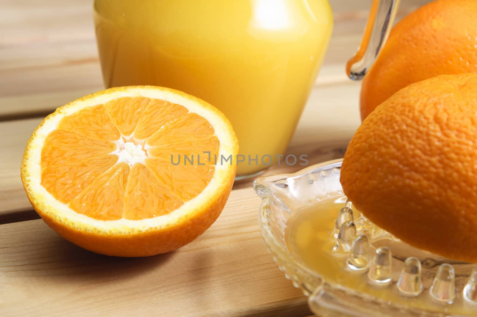 A jug of orange juice with whole and cut oranges and an orange squeezer on a wooden table.