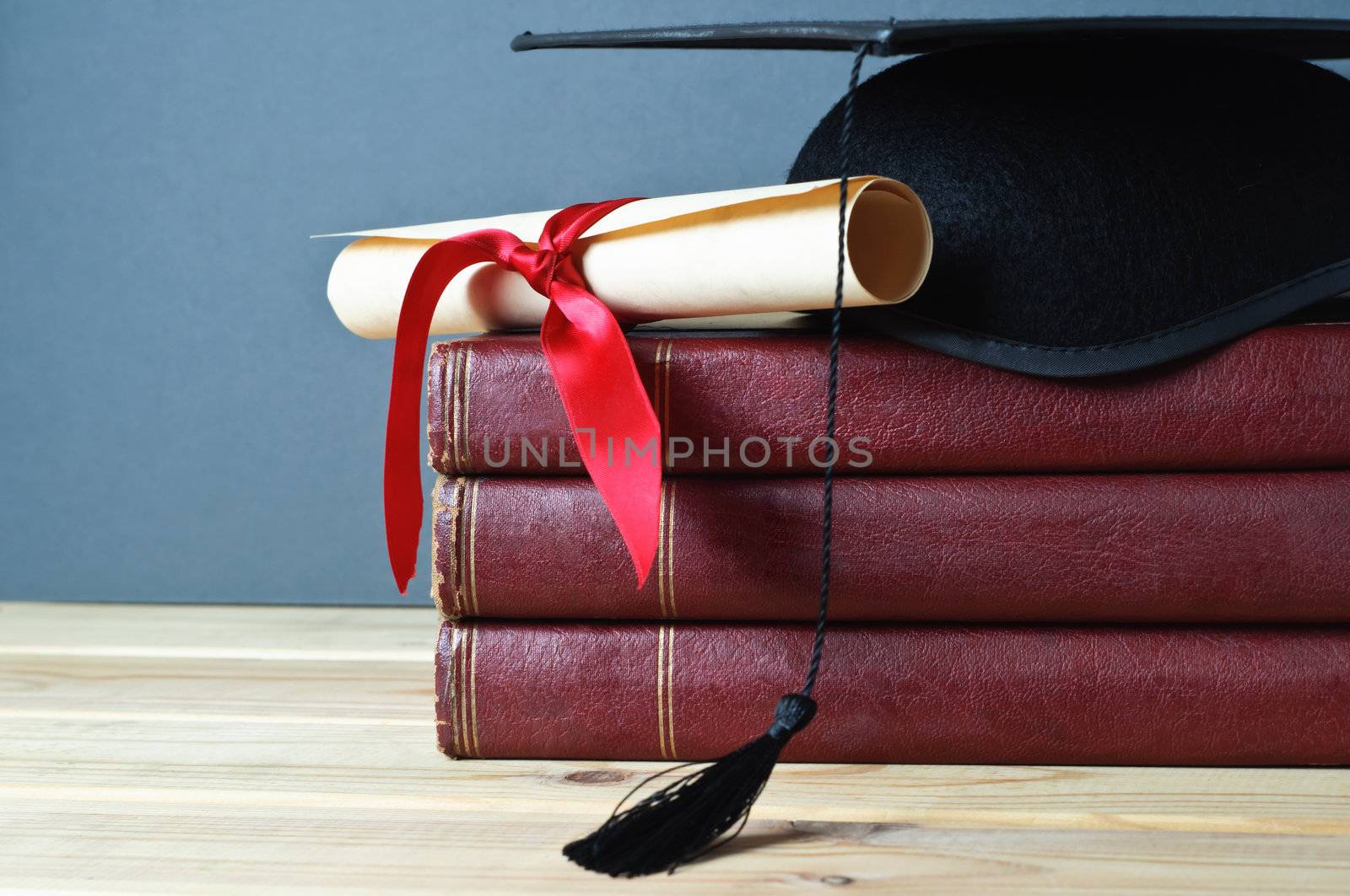 Graduation mortarboard and scroll tied with red ribbon on top of a stack of old, worn books on a light wood table.  Grey background.  