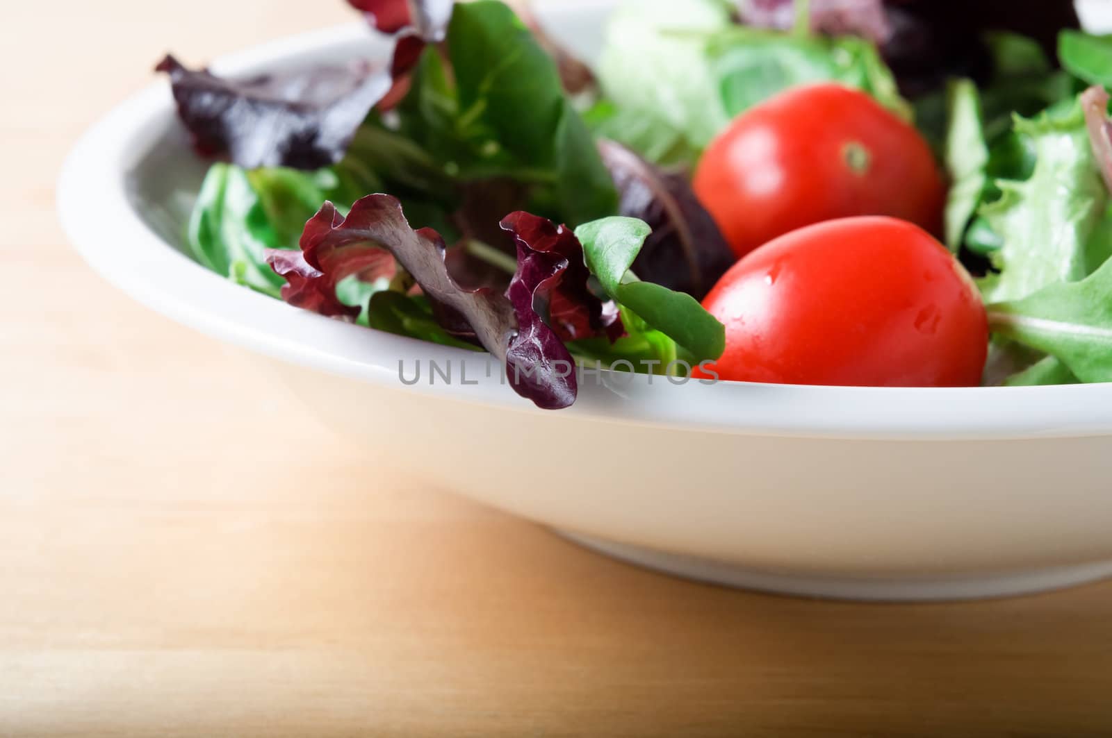 A white bowl of salad, including red lettuce, rocket and tomatoes, on a light wooden table.  Landscape (horizontal) orientation.