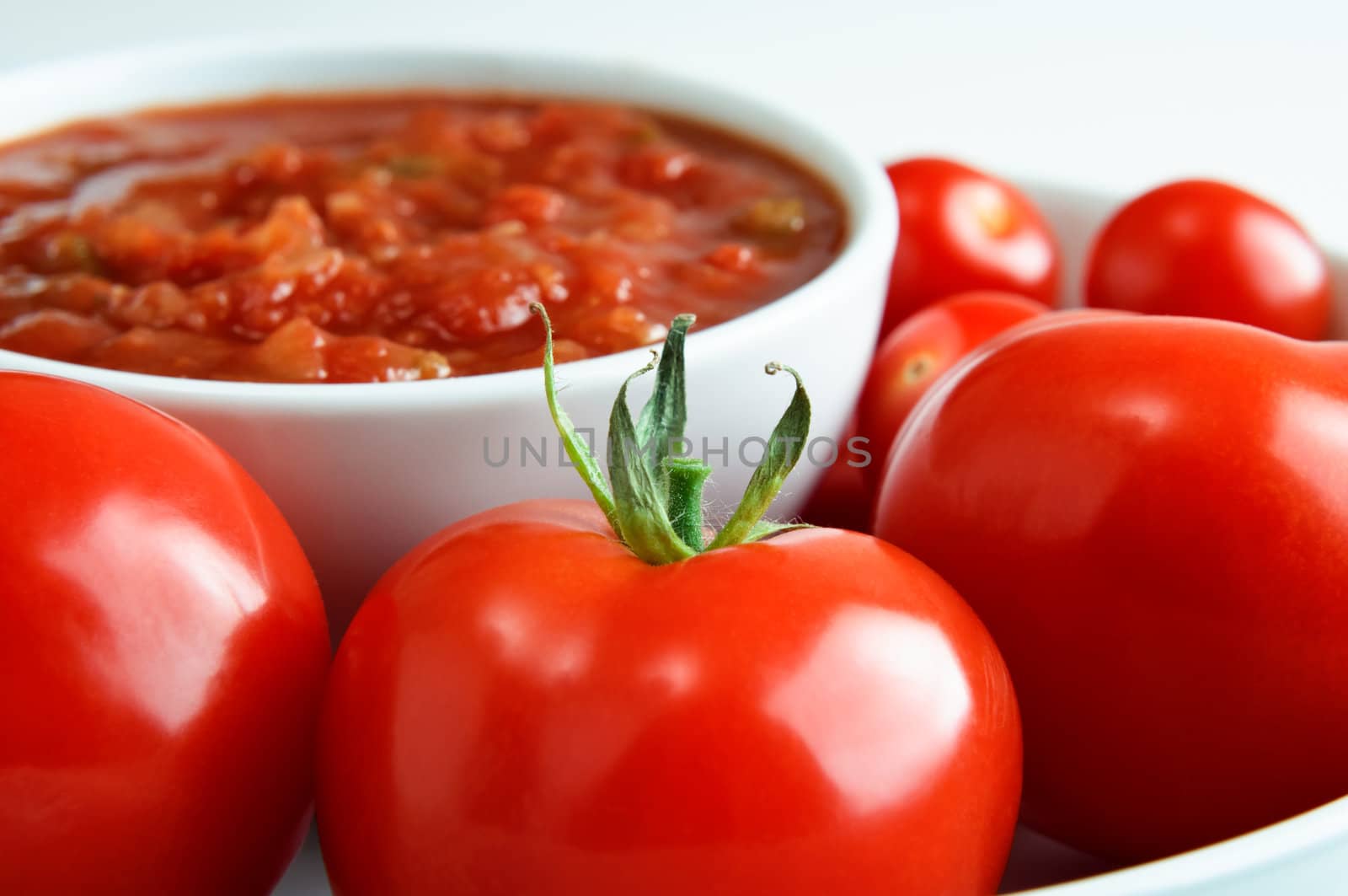 Close up (macro) of bright red tomatoes, surrounding a bowl of salsa in soft focus in the background.  Horizontal (landscape) orientation.