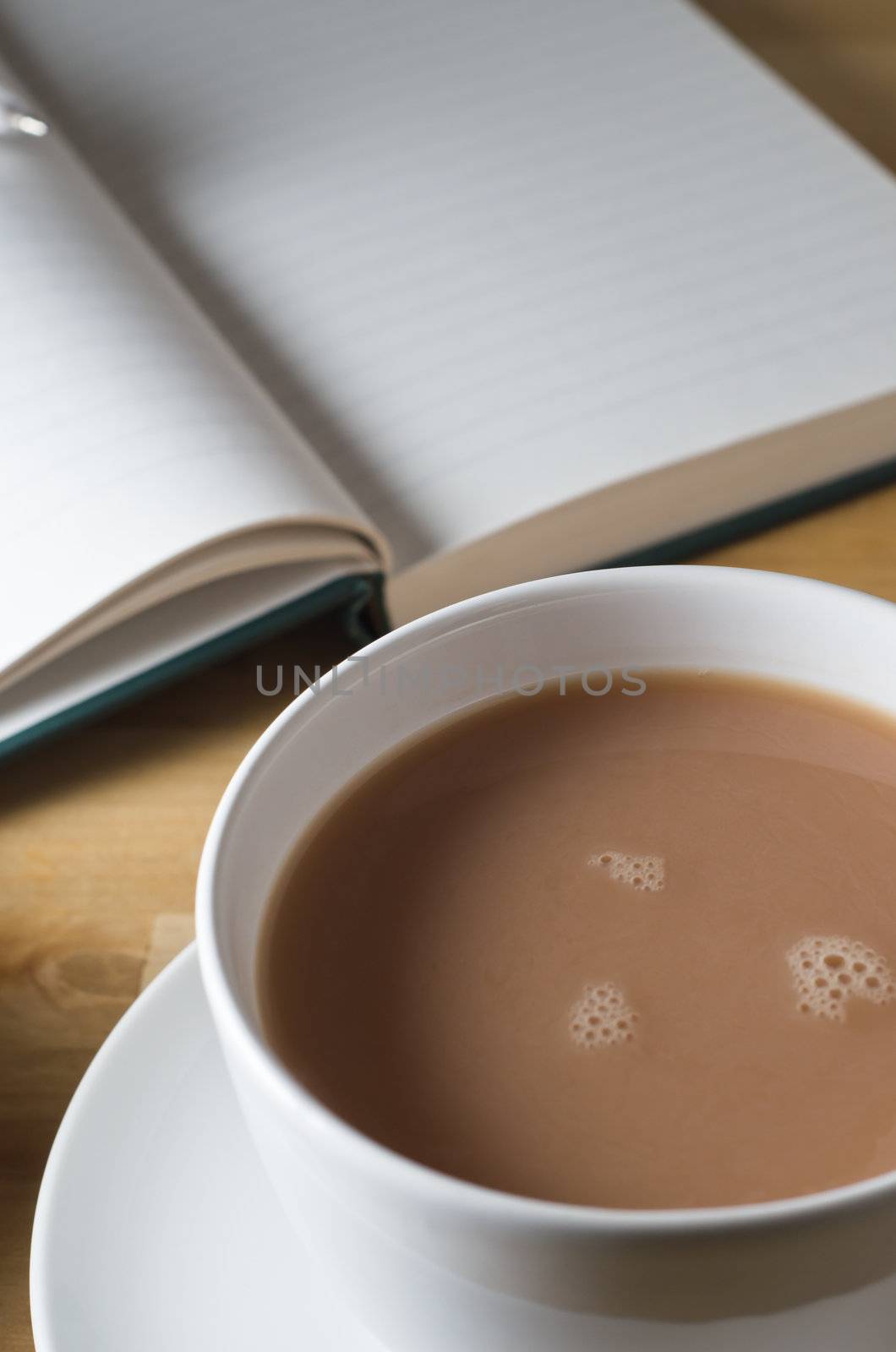 Portrait (vertical) close up of a cup and saucer of tea on a wooden table, with a journal an open, blank journal and pen in soft focus background. 