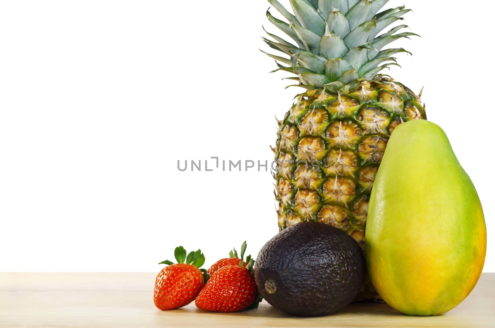 A fruit composition  of pinapple, mango, avocado and strawberries on a light wooden table against a white background.  Copy space to left.