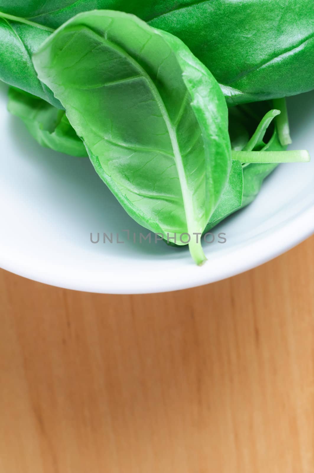 Close up (macro) of whole green basil leaves in a white china bowl, on a light wooden surface. Vertical orientation with copy space below.