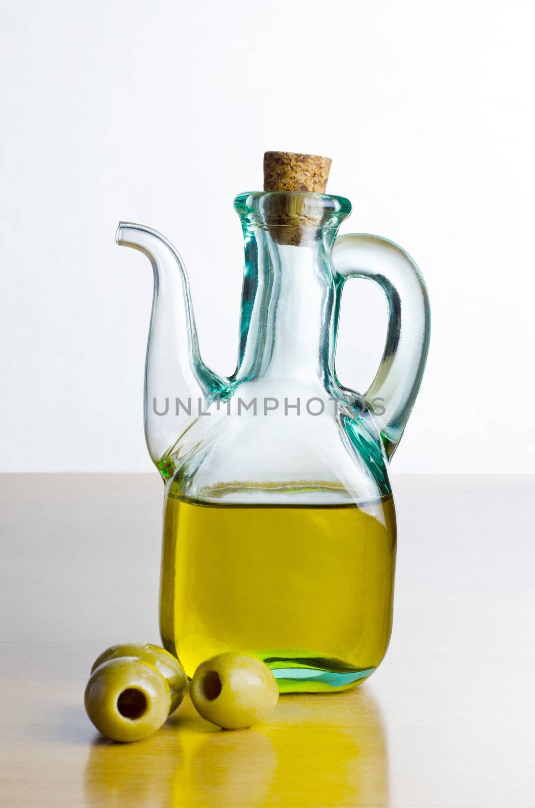 A corked glass jug of olive oil, with pitted olives on a shiny wooden surface with an off-white background.