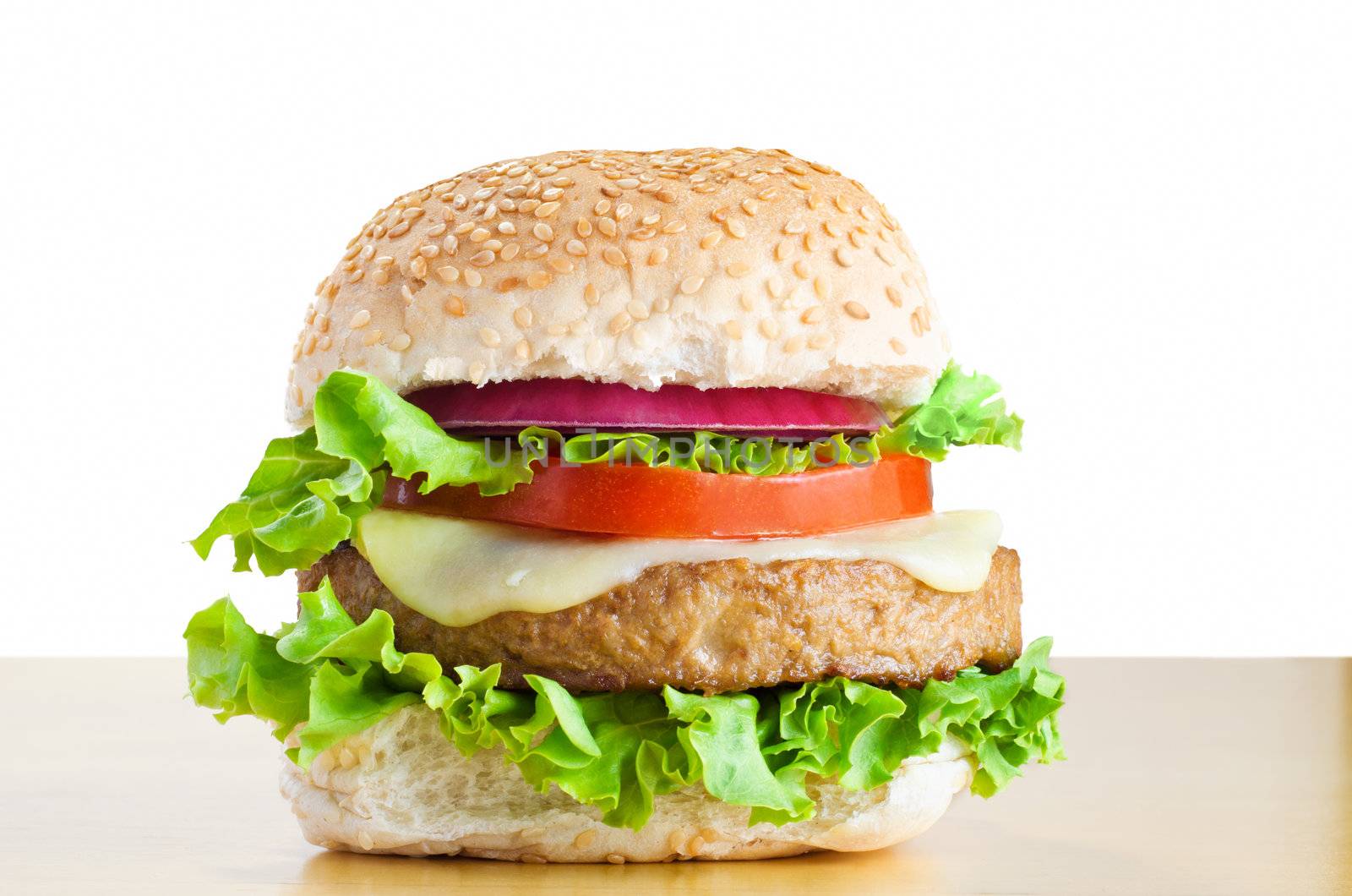 A veggie burger (made from soya protein) in a sesame seed bap with layers of curly lettuce, melted cheese, tomato and onion, on a light wood table with white background.