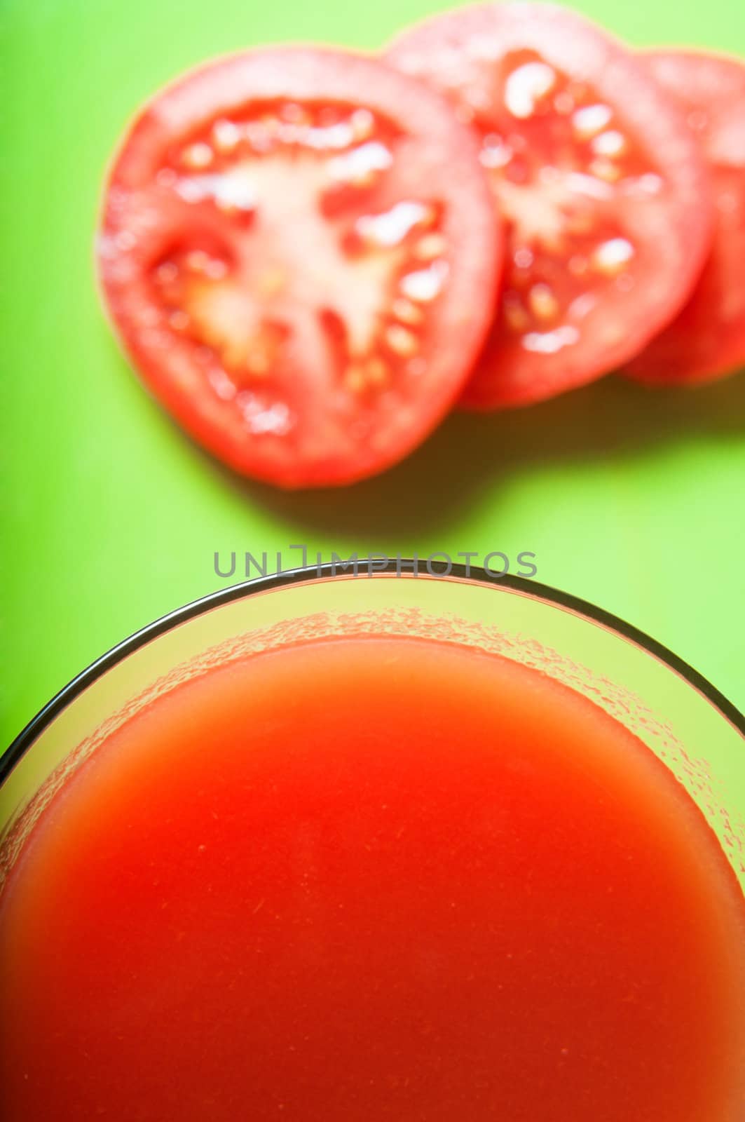 Overhead close up of a glass of tomato juice with tomato slices in soft focus background on green surface.  