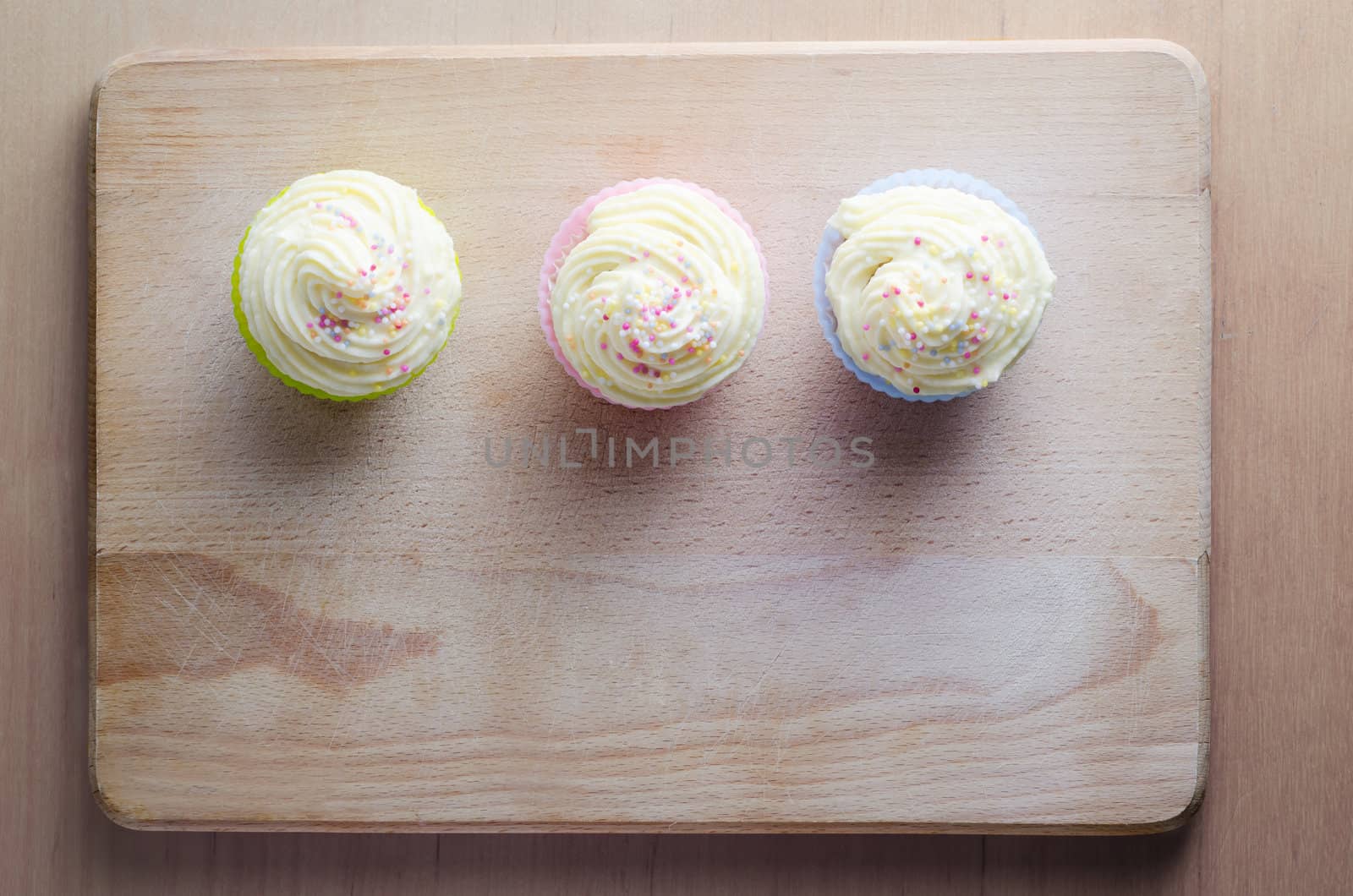 Overhead shot of three cupcakes on a wooden chopping board, topped with swirls of buttercream icing and colourful sprinkles.