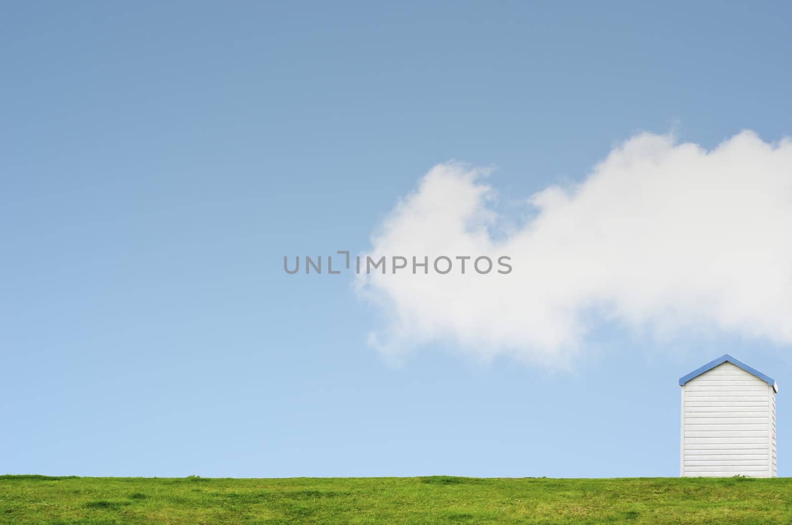 A solitary beach hut in lower right of frame on a green grassy hill against a bright blue sky with a big white fluffy cloud. 