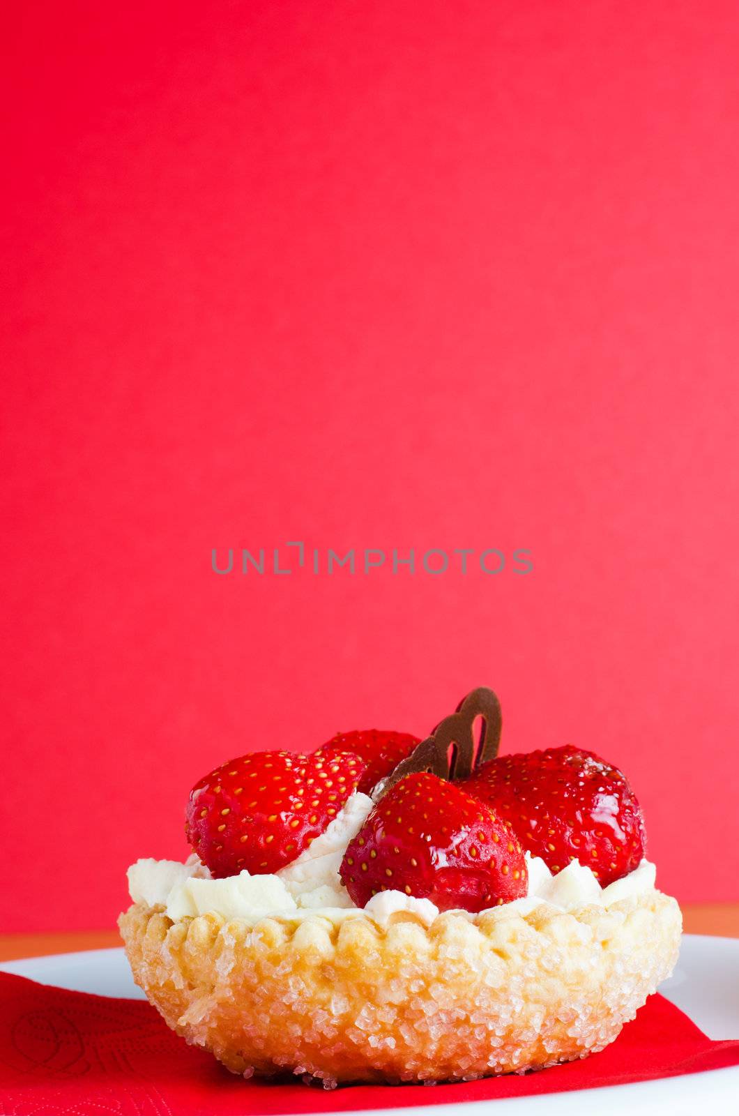 A strawberry and cream pastry cake with chocolate decoration on red napkin and white plate, against a red background.