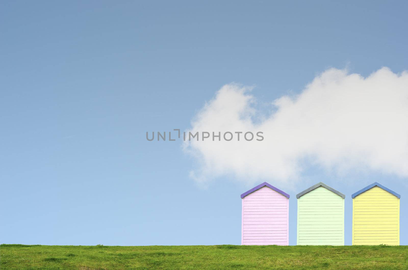 A row of three colourful beach huts on top of a grassy hill against a bright blue sky with white fluffy cloud. Copy space above and left.