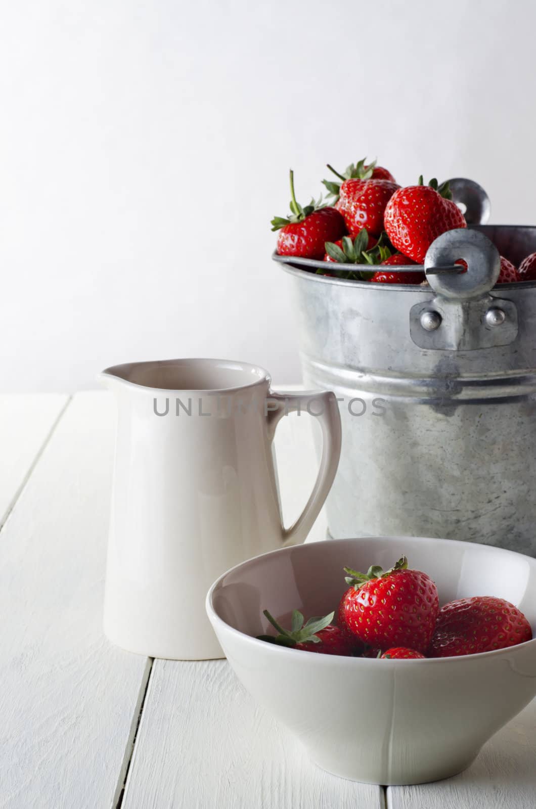 A rustic table scene of freshly harvested strawberries, just harvested.  Includes a tin pail of strawberries, a cream jug and a bowl of served strawberries. 