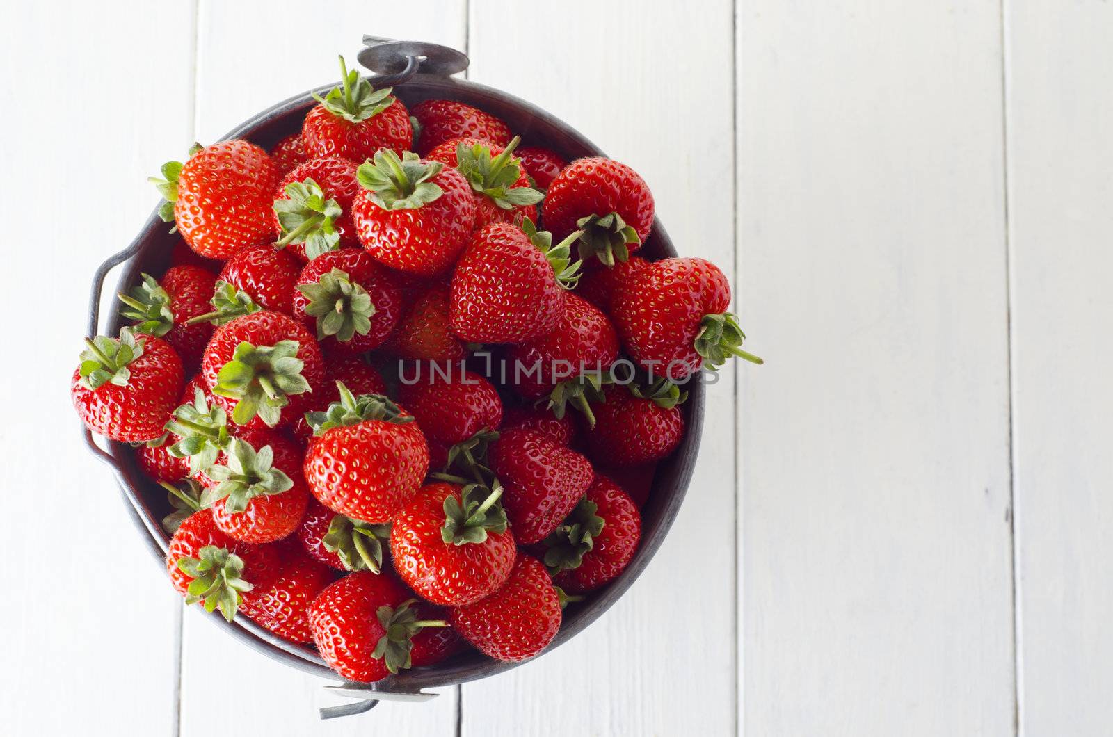 Overhead shot of a tin pail filled to the top with fresh strawberries, on a wooden planked table, painted white.  Copy space to right of frame.