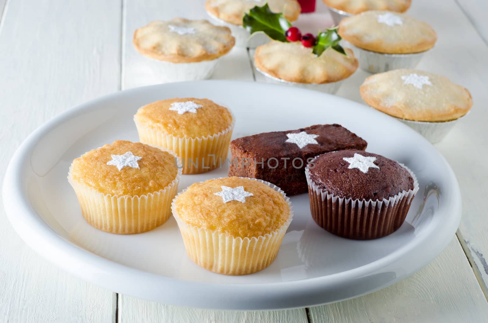 Christmas cakes on a plate decorated with stars from sifted icing sugar, with mince pies and holly in soft focus background. Laid out on a white, planked farmhouse table.