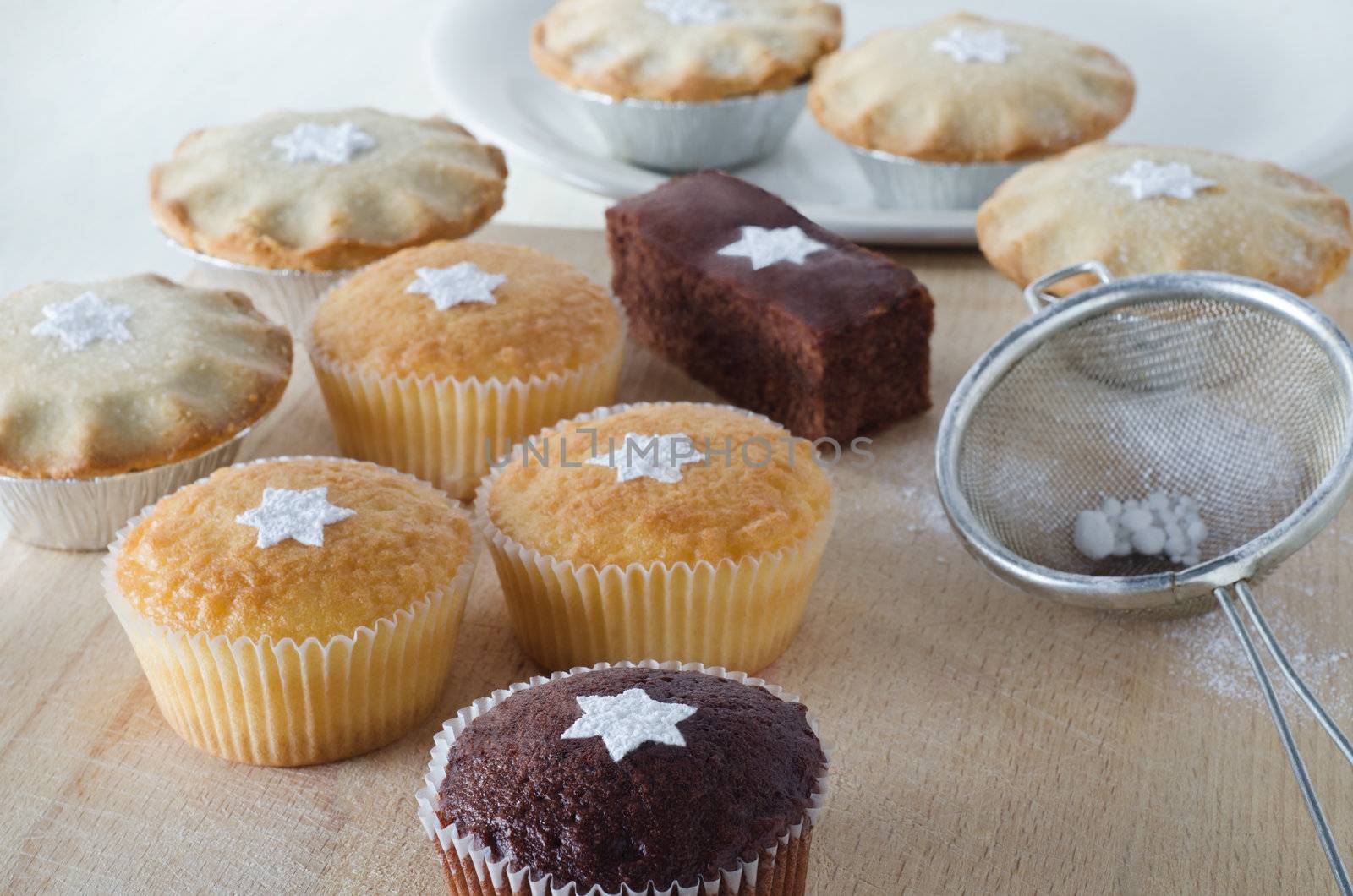 A group of Christmas cakes and mince pies, decorated with star shapes from sifted icing sugar, on a wooden chopping board with plate in background, and sifter with scattered icing sugar to the right. 