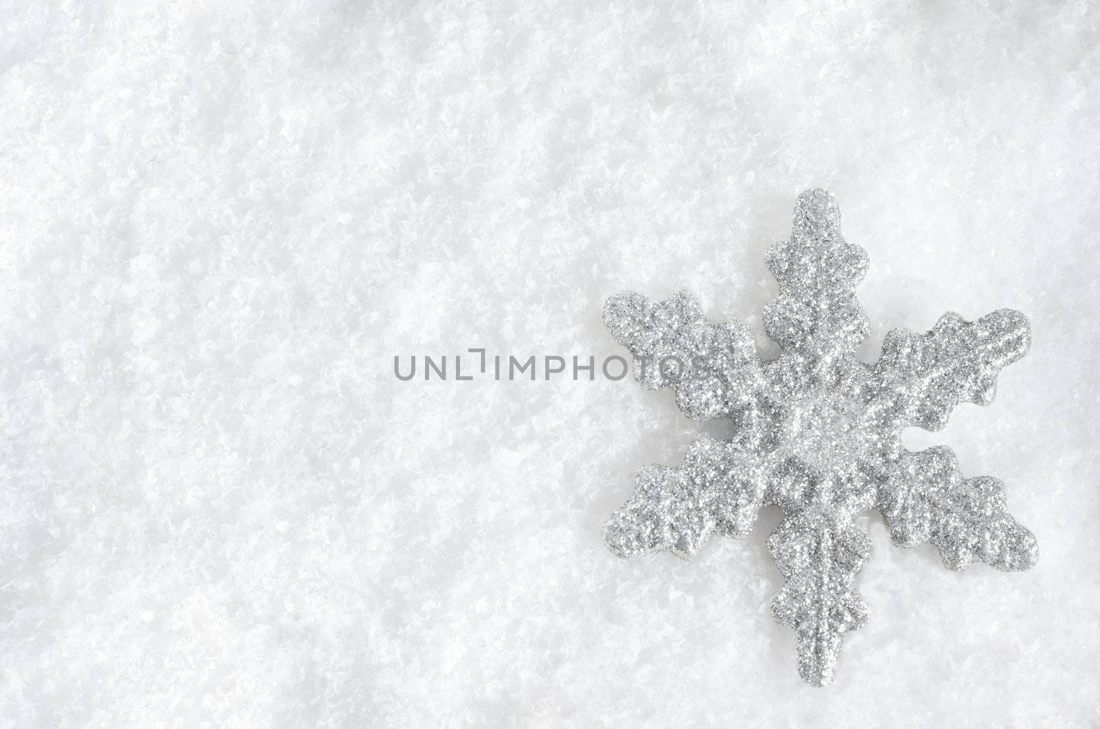 Winter background shot from above.  Glittery silver Christmas snowflake faces upwards on lower right, lying in fake snow.