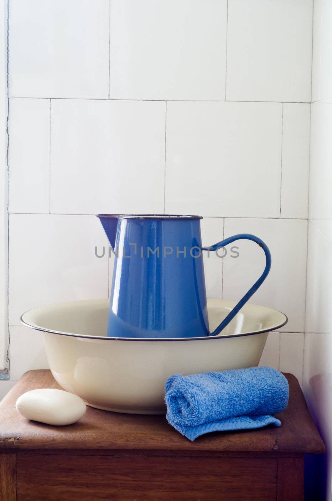 Rustic, retro bathroom washstand scene comprising an enamal jug inside a bowl with rolled flannel and soap on an old wooden cabinet, lit by natural window light from the left.   Shabby white, tiled wall in the background provides copy space.