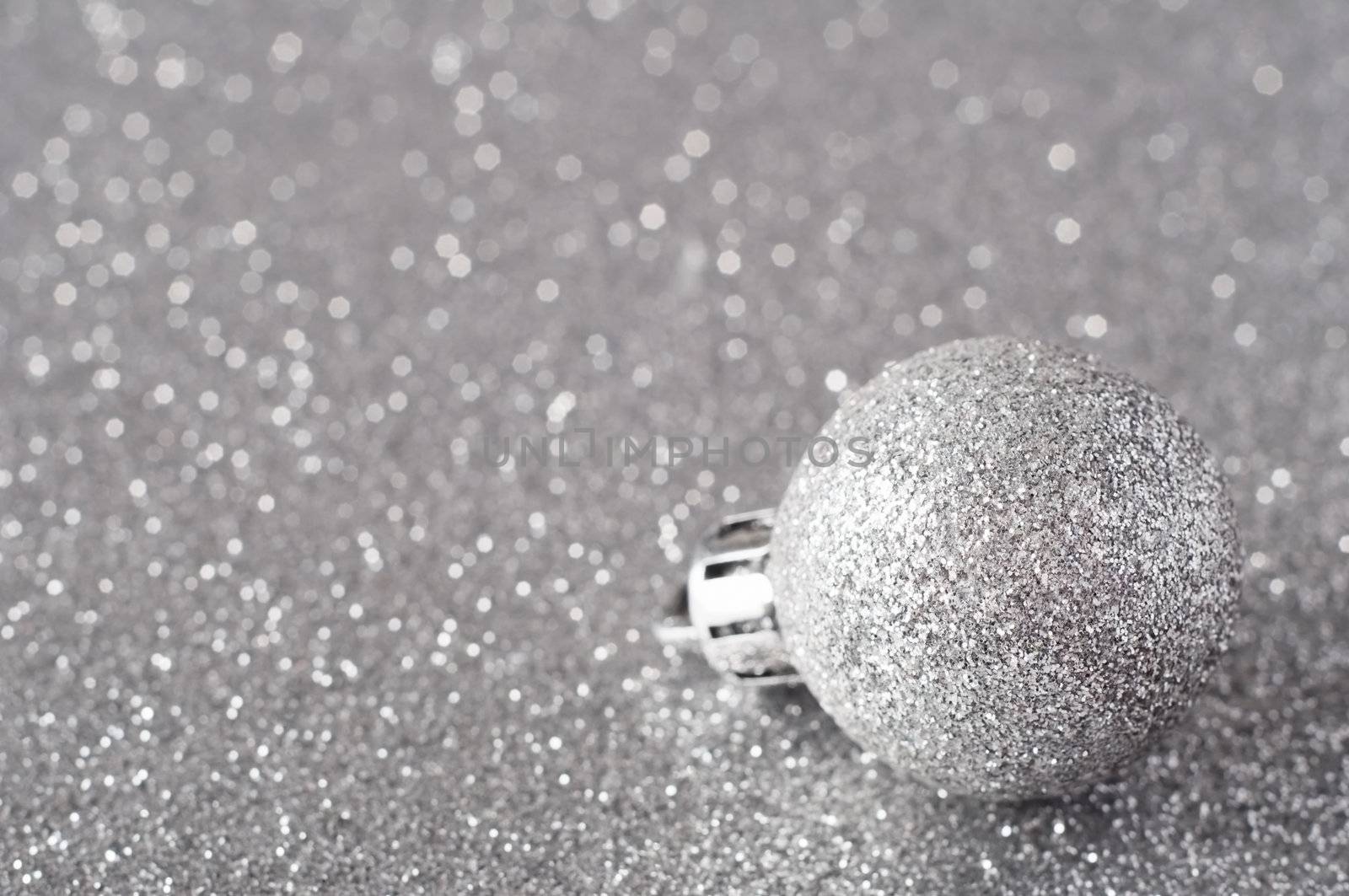 A single silver bauble, coated in glitter, resting on a silver glitter surface that softens into bokeh in the background.