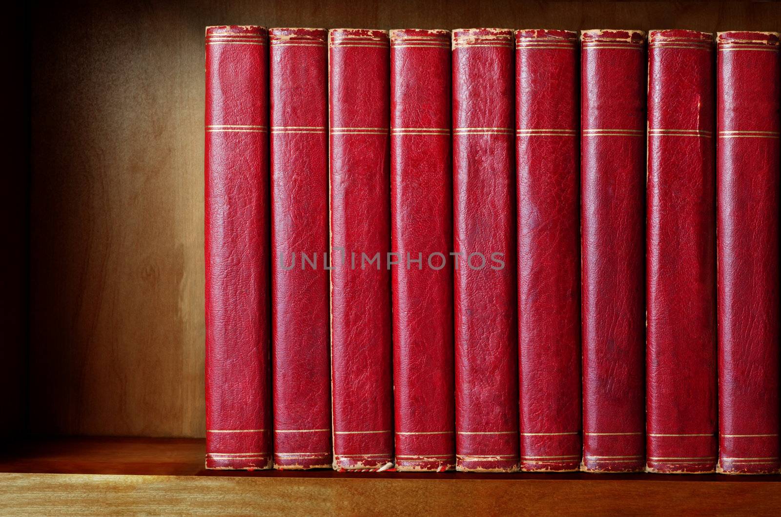 A row of old, battered, matching encyclopaedias (circa 1950s) lined up on a shelf, with titles removed to leave blank spines.  Red leather effect with gold striped trims.  Shelf has been darkened artificially to give impression of age.