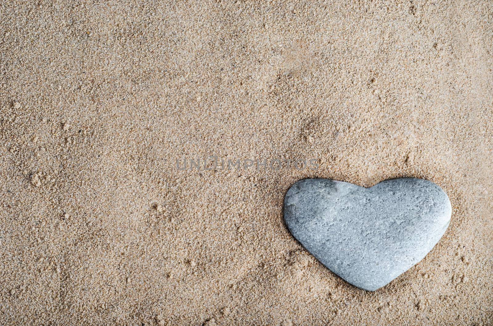 A grey heart shaped stone nestled in golden sand at bottom right of frame.  Copy space in sand.