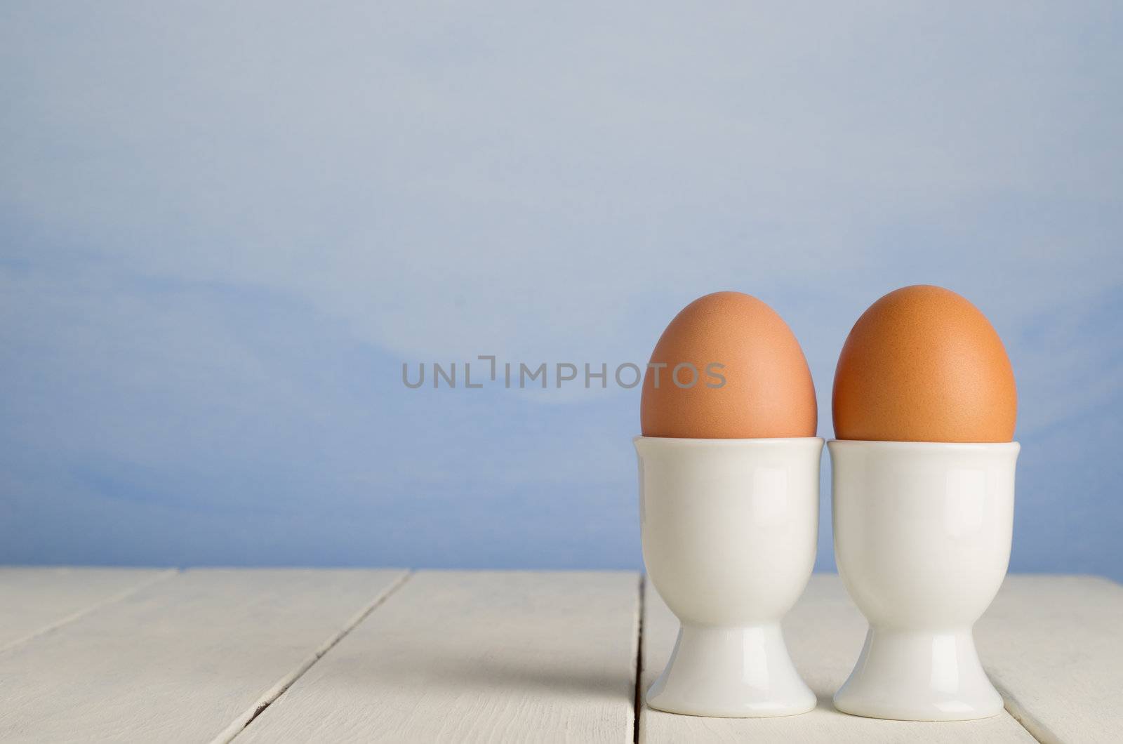 Two fresh brown undecorated eggs, side by side in white porcelain egg cups against a painted blue sky effect background, on an old cream painted wood plank farmhouse table.
