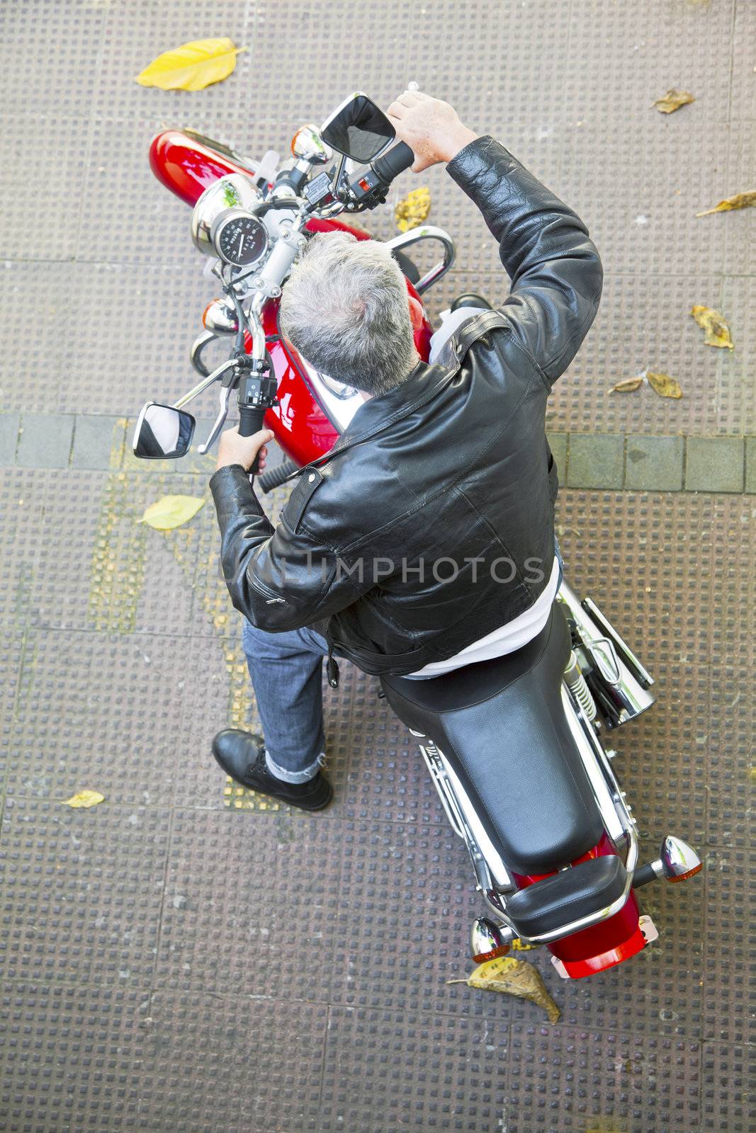 Diagonal aerial viewpoint of a stationary red motorbike and a Grey haired senior rider in leather jacket, denim jeans and boots on a tied floor with fallen autumn leaves. Generic shot taken in Bombay India