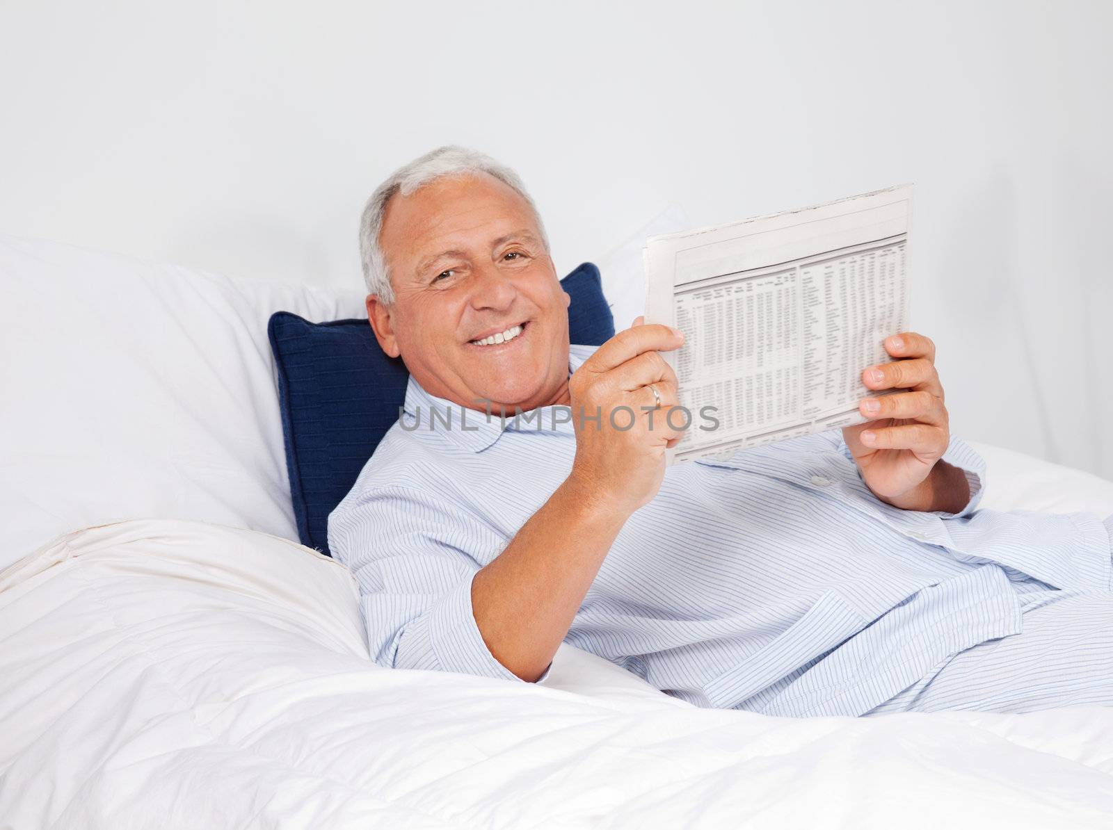 Portrait of relaxed senior man reading newspaper while lying on bed at home