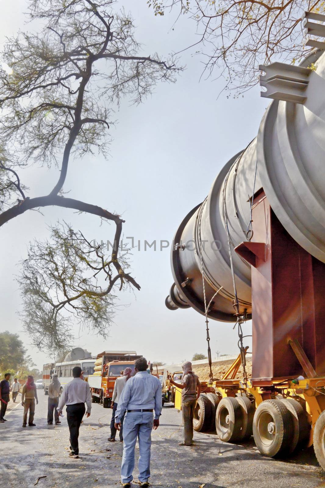 A heavy goods transporter has his cargo stuck between two trees on either side of a tree lined road and road crew and emergency services clearing the accident and traffic jam on 25th February 2013, Vertical landscape caught on the coastal road in Gujarat India, between Bhavnagar and Mangrol.