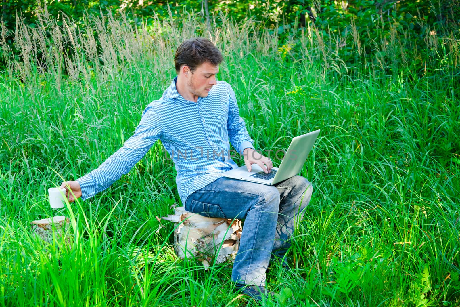 Young man outdoors with a laptop reaching his coffee