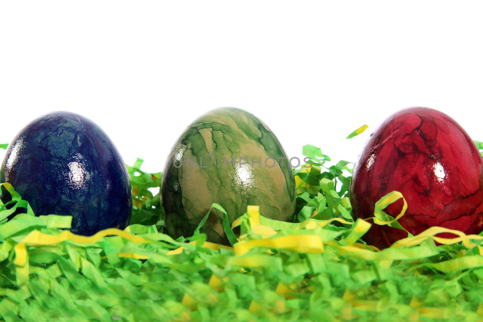 Three marble patterned Easter Eggs in blue, green and red nestled in a bed of green artificial straw against a white background