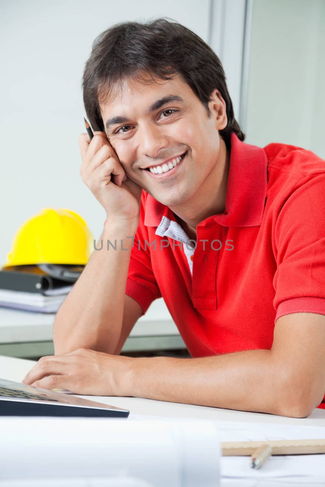 Portrait of young male architect smiling while sitting by desk with laptop