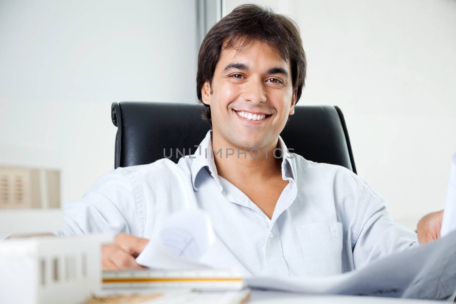 Portrait of happy young male architect with blueprint sitting in office chair