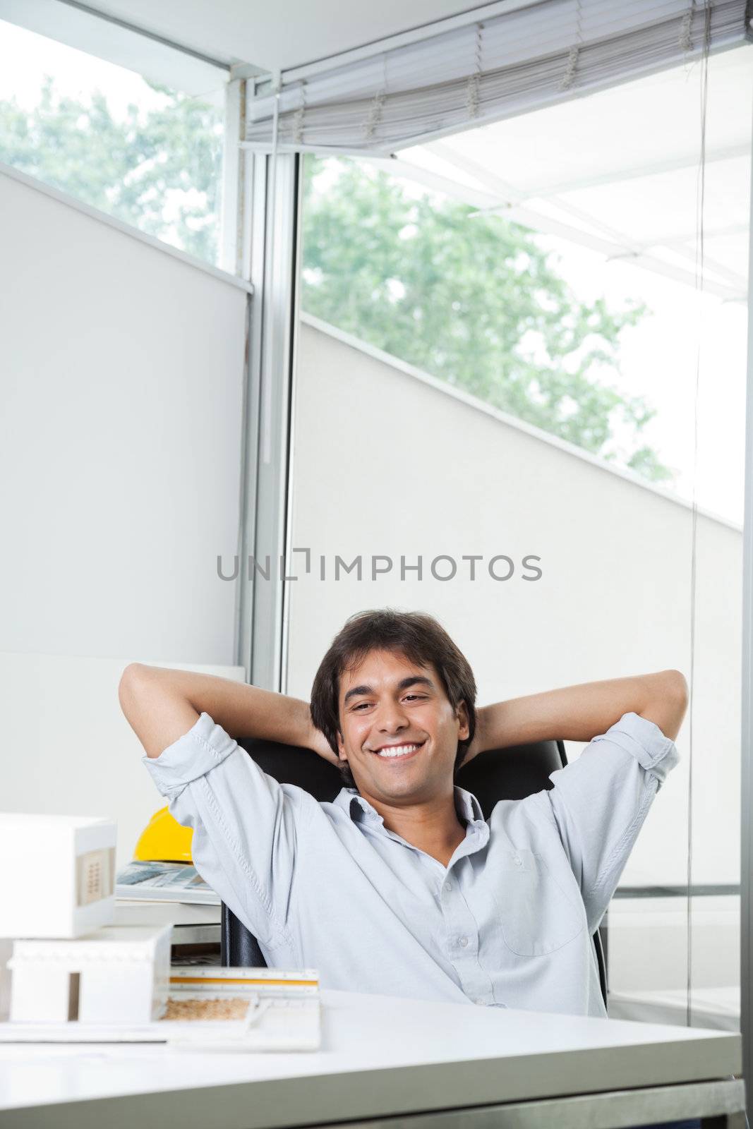 Happy young male architect looking at model house while sitting in chair with hands behind head